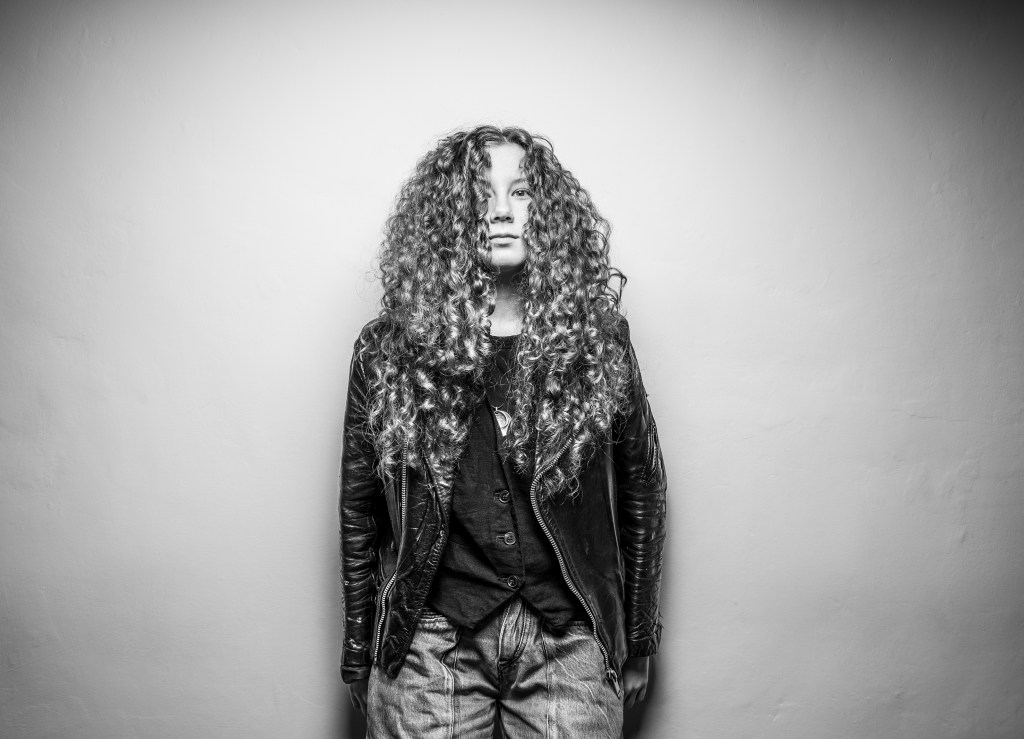 A portrait of a person with long, curly hair wearing a black leather jacket and casual jeans, standing against a plain wall.
