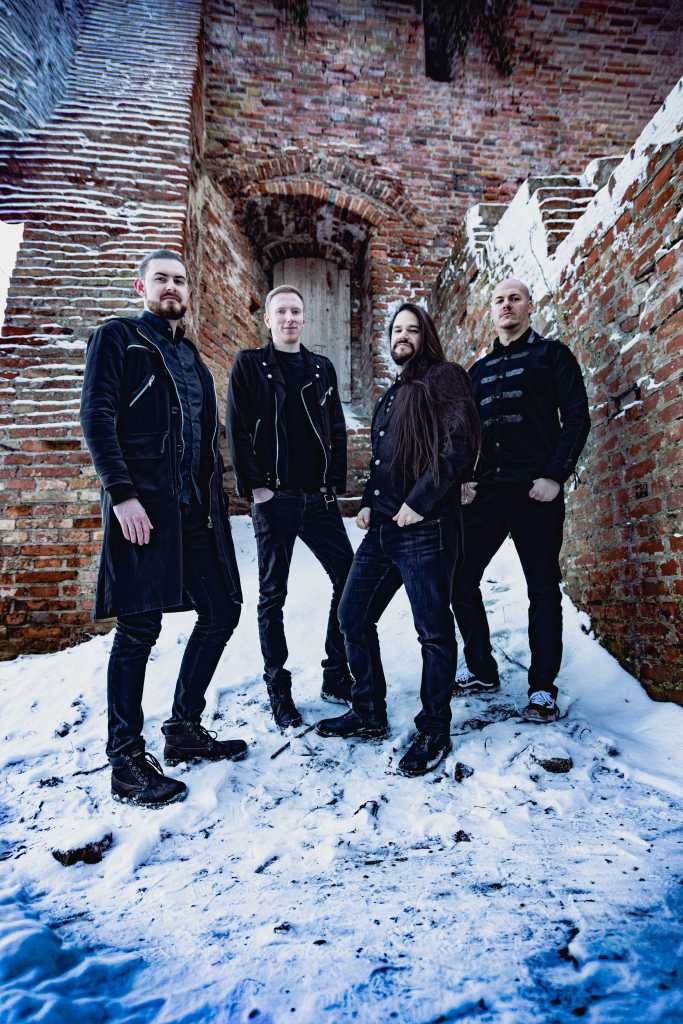 A band of four male musicians posing in front of a rustic brick wall with a snowy ground, wearing dark, stylish clothing.