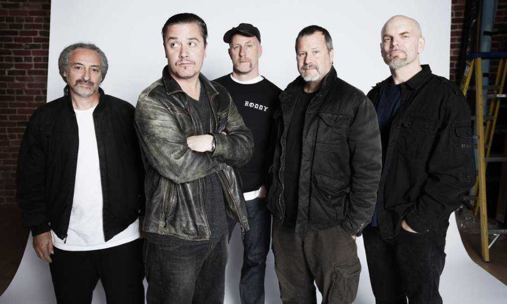 A group of five men posing together in front of a plain white backdrop, wearing a mix of casual and stylish clothing, with expressions ranging from serious to relaxed.