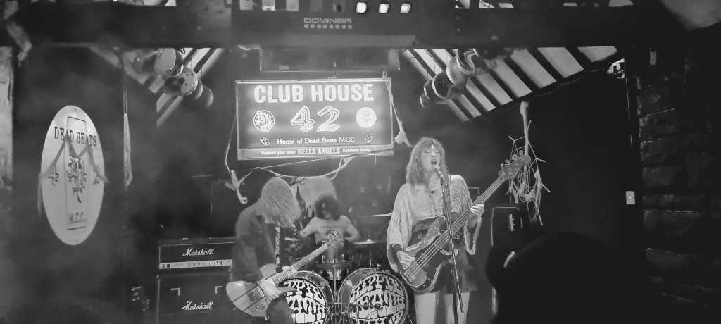A black and white image of a live music performance at Club House 42, featuring three musicians on stage. One musician plays the bass guitar, another is on the drums, and the third plays an electric guitar. The stage is decorated with band posters and the venue has a rustic appearance.