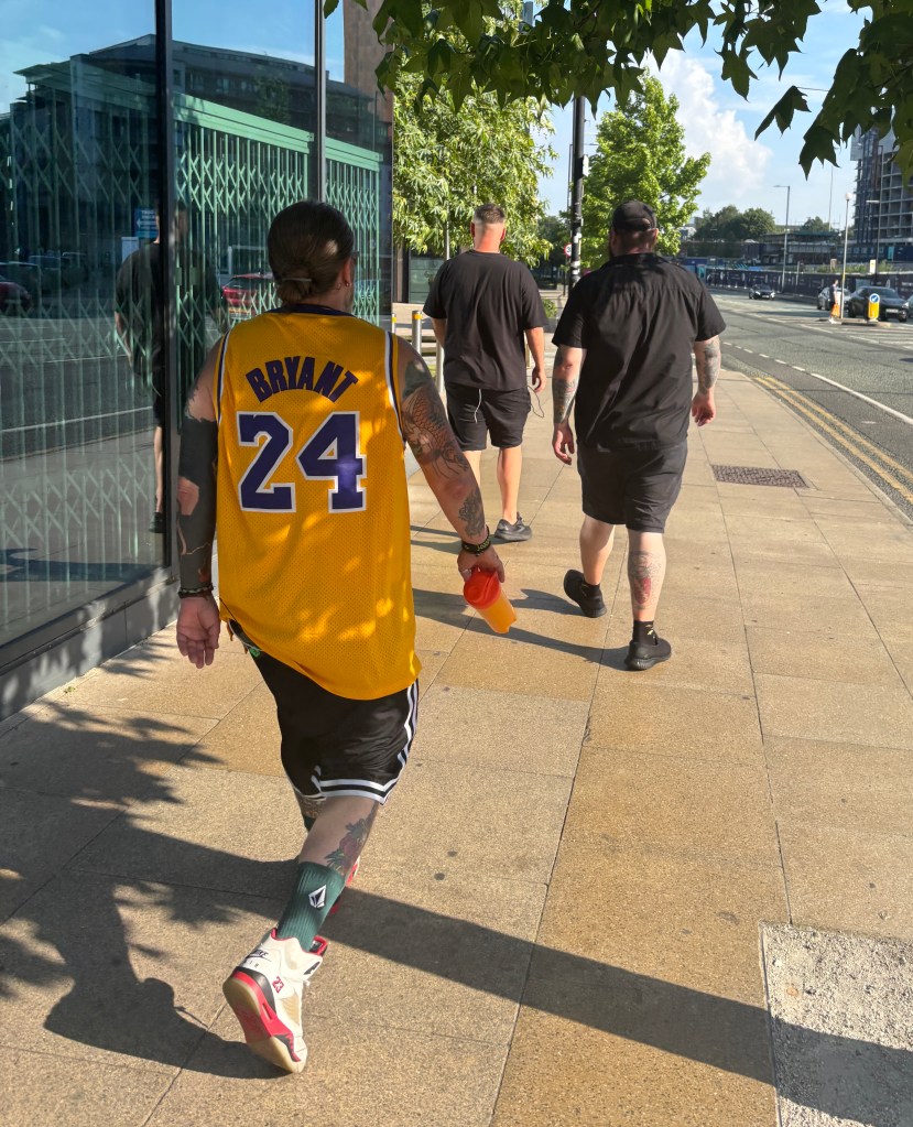 A group of three individuals walking on a sunny day, with one person wearing a yellow basketball jersey featuring the name 'Bryant' and the number 24. The background includes buildings and greenery.